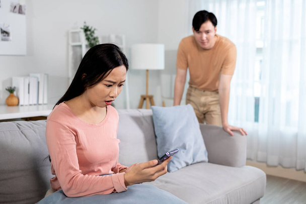 Shocked jealous Asian girlfriend suspecting infidelity by reading text messages on her cheating boyfriends phone at home. Wife checking husbands smartphone and having jealousy problem - Photo, image