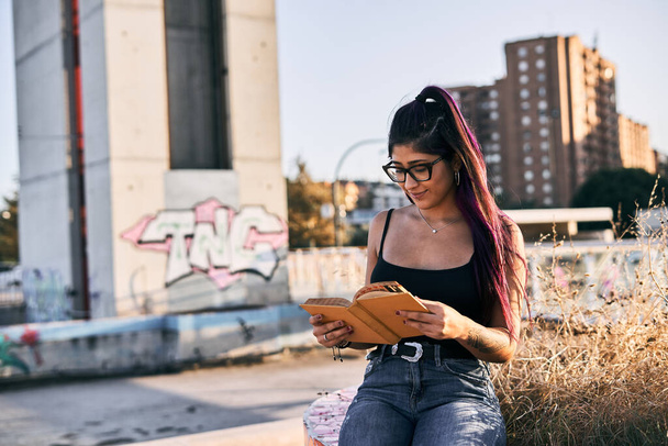 menina latina com óculos morena colar de cabelo longo em seu pescoço piercing no lábio sorrindo enquanto lê um livro no parque da cidade - Conceito urbano - Foto, Imagem