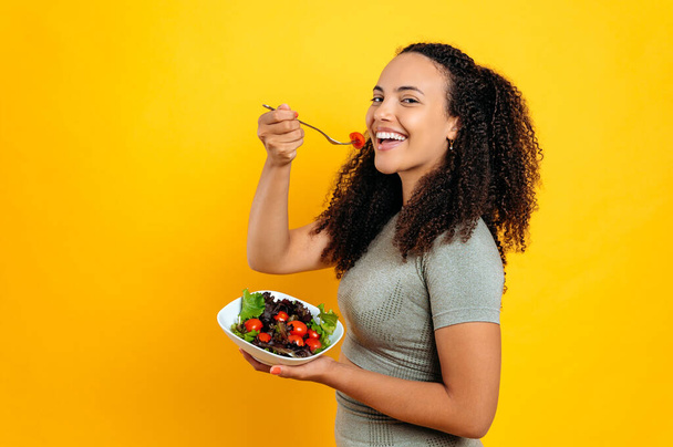 Sağlıklı yaşam tarzı, vejetaryen. Güzel, melez, kıvırcık saçlı spor elbiseli kadın elinde domatesli taze salata, izole edilmiş turuncu arka planda duruyor sebze yiyor, gülümsüyor. - Fotoğraf, Görsel