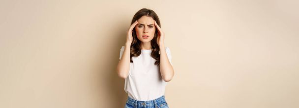 Health and women concept. Portrait of girl feeling sick, touching head temples, feeling headache, migraine, standing over beige background. - Foto, Imagem