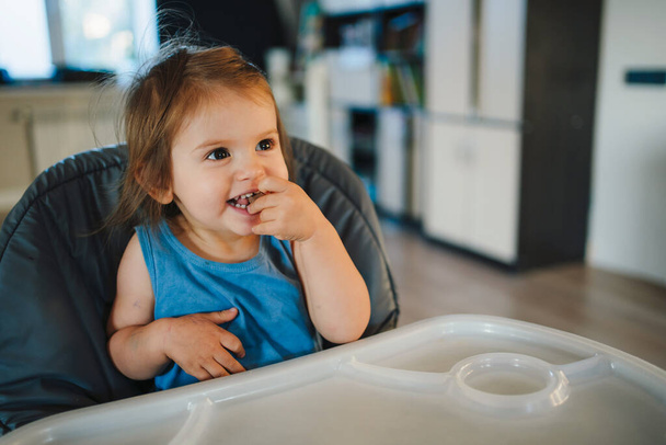 Menina adorável sentada em cadeira alta, comendo um pedaço de chocolate em casa. Retrato de close-up das crianças. Conceito de estilo de vida bebê - Foto, Imagem
