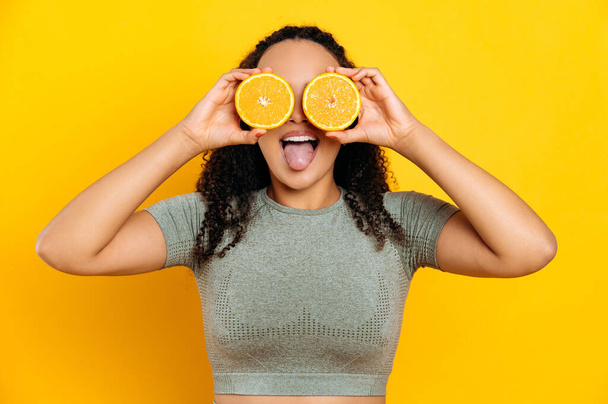 Радісна красива бразильська або латиноамериканська молода кучерява жінка, в спортивних костюмах, стоїть на ізольованому помаранчевому фоні, тримає дві половинки апельсина в руках біля очей, показує язик, посміхається
 - Фото, зображення