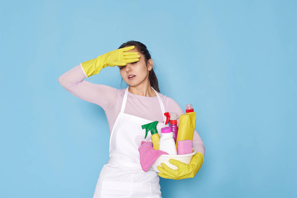 tired caucasian woman in gloves holding bucket with cleaning supplies on blue background. - Foto, Imagem