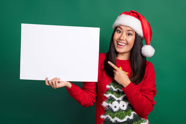 Photo of pretty sweet girl dressed ugly print red x-mas pullover headwear pointing white billboard empty space isolated green color background. - Photo, image