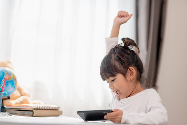 Aasialainen pikkutyttö käyttää puhelinta lähikuva, hajamielinen opiskelusta, istuu pöydässä muistikirjojen kanssa, kaunis lapsi pitää hauskaa älypuhelimen kanssa, katselee webinaaria, kotiopetusta - Valokuva, kuva