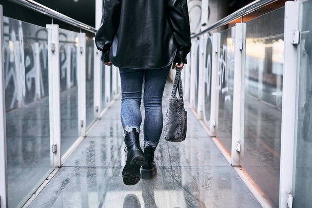 parte posterior de las piernas de una chica latina en pantalones azules chaqueta de piel y grandes botas negras caminando por la pasarela de cristal - Concepto urbano - Foto, Imagen