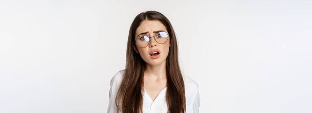 Großaufnahme einer verwirrten brünetten Frau mit Brille, die verwirrt und ratlos vor weißem Hintergrund steht. Kopierraum - Foto, Bild