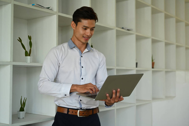 Αυτοπεποίθηση ασιάτης επιχειρηματίας κοιτάζοντας φορητό υπολογιστή, διαβάζοντας τα μηνύματα ηλεκτρονικού ταχυδρομείου των επιχειρήσεων, ενώ στέκεται στο σύγχρονο γραφείο. - Φωτογραφία, εικόνα
