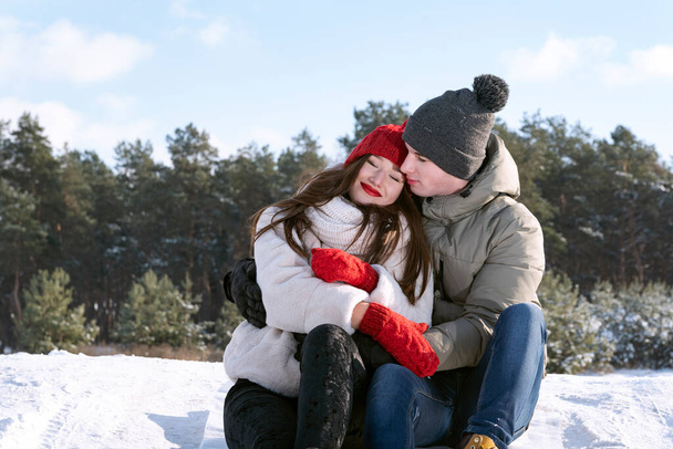 Jongeman en vrouw zitten en knuffelen tegen de achtergrond van het winterbos. Zonnige winterdag. - Foto, afbeelding