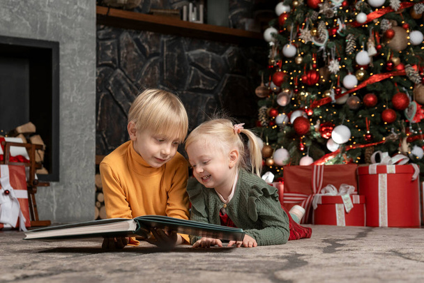 Deux frères et sœurs souriants reposent par terre sous le sapin de Noël. Frère et sœur blonds lisent le livre. Veille de Noël. - Photo, image