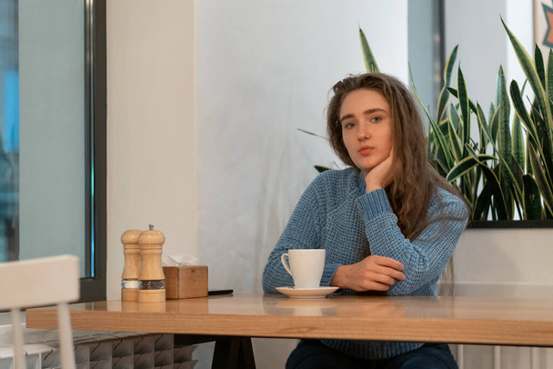 Menina bonito com cabelo castanho usa camisola de malha azul senta-se no café com xícara de chá de café, cappuccino latte e olha atenciosamente para a câmera - Foto, Imagem