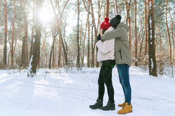 Młoda szczęśliwa para w słoneczny dzień w ośnieżonym parku. Mężczyźni i kobiety stoją i przytulają się w śnieżnym zimowym parku. Widok boczny. - Zdjęcie, obraz