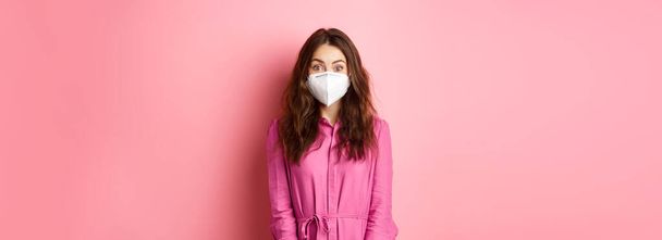 Covid-19, pandemic and lifestyle concept. Excited young woman wearing respirator while going out, social distance herself during quarantine, pink background. - Photo, Image