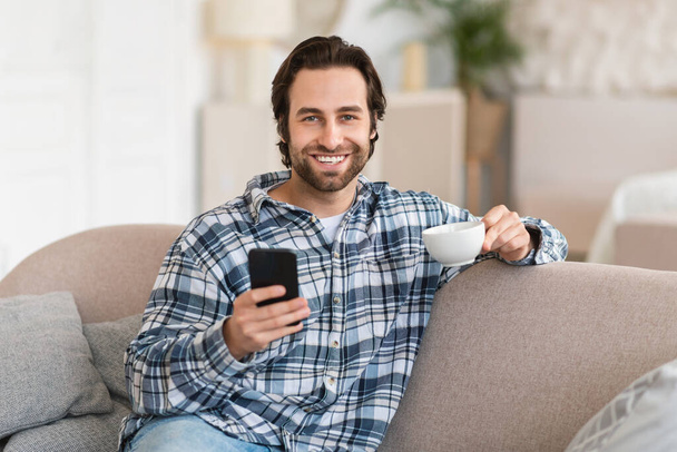 Счастливый тысячелетний европеец в домашней одежде с щетиной на смартфоне, смотрит видео, наслаждается чашечкой горячего напитка в гостиной. Приложение для социальных сетей, блог и кофе-брейк дома - Фото, изображение