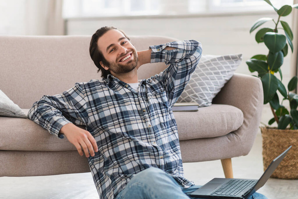 Happy millennial european male in domestic clothes with stubble sits on floor, working on computer in living room interior, copy space. Break, rest, relaxation from job and business, lifestyle at home - Valokuva, kuva