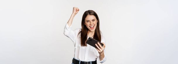 Happy brunette woman playing mobile video game, smiling and looking at screen excited, standing over white background. - 写真・画像