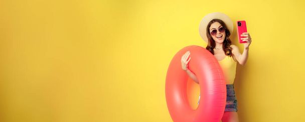 Stylish brunette girl on vacation, taking selfie with swim ring, going on beach, swimming in sea on summer holiday, standing over yellow background. - Photo, image