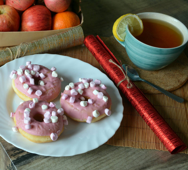 Cup of black tea with lemon and three donuts with pink icing on a plate on a wooden table. Sweet breakfast. - Photo, Image