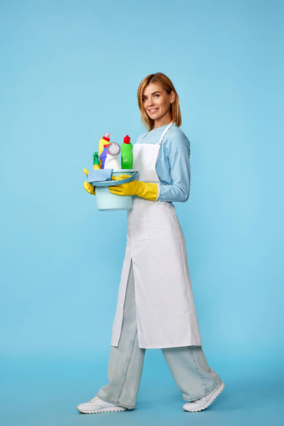 χαρούμενη χαρούμενη ξανθιά γυναίκα με λαστιχένια γάντια και καθαρότερη ποδιά με προμήθειες καθαρισμού σε μπλε φόντο. Πλήρες μήκος - Φωτογραφία, εικόνα