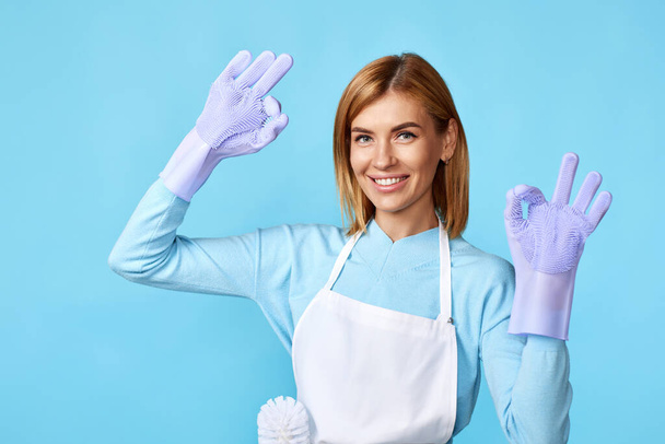 beautiful caucasian blonde woman in violet rubber gloves and cleaner apron showing ok gesture on blue background. cleaning - Fotografie, Obrázek