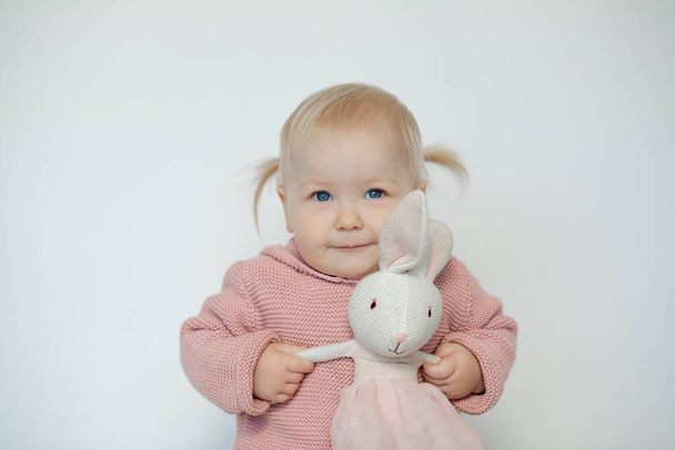 Cute baby girl play with stuffed animal, isolated on white. Smiling toddler gladly holding by hands teddy bunny. Blonde haired child in pink clothes with paschal rabbit. Easter. Holiday gift. - Φωτογραφία, εικόνα