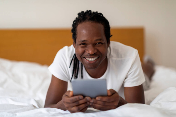 Fröhlicher junger schwarzer Mann mit digitalem Tablet, das im Bett zu Hause ruht, fröhlicher Afroamerikaner mit Dreadlocks-Frisur, der Pad-Computer für Online-Einkäufe oder das Surfen im Internet benutzt, freier Raum - Foto, Bild
