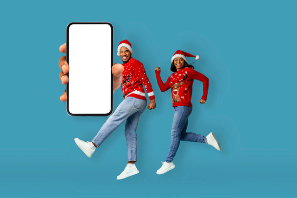 冬の販売をキャッチするために空気中で実行されている鹿とサンタの帽子と赤いクリスマスのセーターで興奮千年アフリカ系アメリカ人のカップル,白い空の画面でスマートフォンを示します,青に隔離されました - 写真・画像