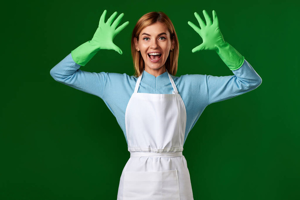 very excited positive woman in rubber gloves and cleaner apron looking at camera on green background. cleaning - Fotó, kép