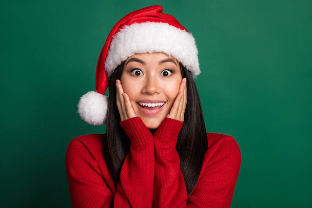 Closeup photo of young korean woman wear red pullover nice santa claus hat touch cheeks shock black friday sale isolated on green color background. - 写真・画像