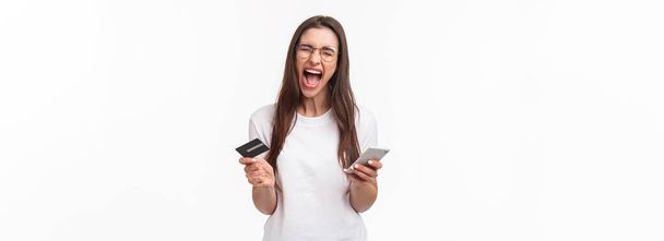 Portrait of uneasy annoyed and angry young brunette female in t-shirt yelling pissed-off as checking bank account, have no money on credit card, or it expired, hold mobile phone, white background. - Fotó, kép