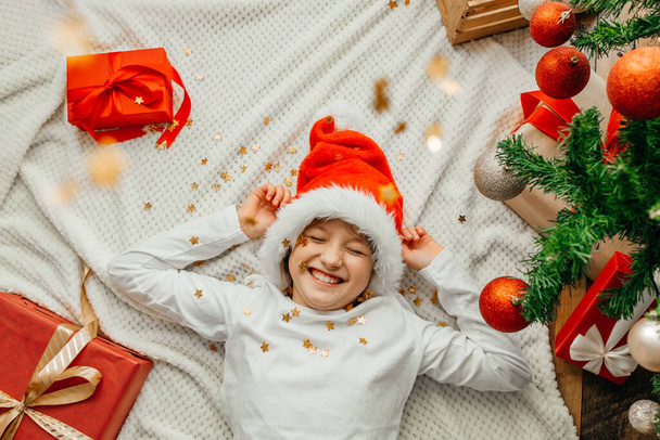 Щаслива дівчина-підліток лежить поруч з новорічною ялинкою, всі посипані золотим конфетті і блискітками
 - Фото, зображення