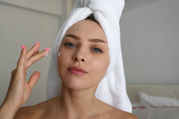 Portrait of young beautiful woman at home performing cosmetic procedures after bathing. Female wearing hair towel wrap applying moisturizing face cream. Close up, copy space, background. - 写真・画像