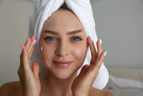 Portrait of young beautiful woman at home performing cosmetic procedures after bathing. Female wearing hair towel wrap applying moisturizing face cream. Close up, copy space, background. - Photo, image