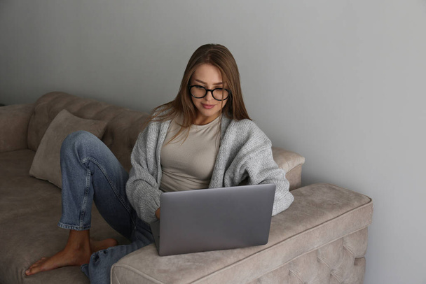 Close up shot of young smiling woman working remotely from home on laptop, sitting on the couch in living room. Female freelancer in blue light glasses with hands on keyboard. Copy space, background. - Fotoğraf, Görsel