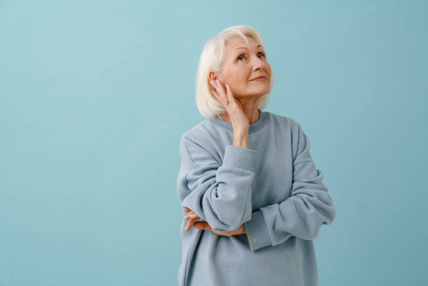 Beautiful thoughtful senior woman in blue pullover looking upward over isolated blue background - Foto, Imagem