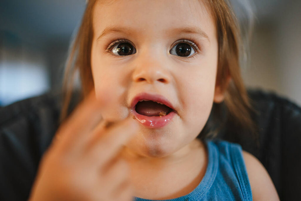 Közelkép egy nagy barna szemű lányról, aki a kezével eszik. Gyönyörű portré baba etetőszék eszik életmód tervezés. - Fotó, kép