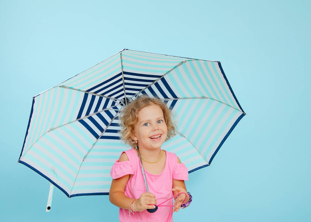 Veselá holčička stojí pod otevřeným deštníkem na prázdném modrém pozadí, volné místo na kopírování. Šťastné dítě školního věku se ukrývá před deštěm. Životní pojištění a bezpečnostní koncepce. - Fotografie, Obrázek