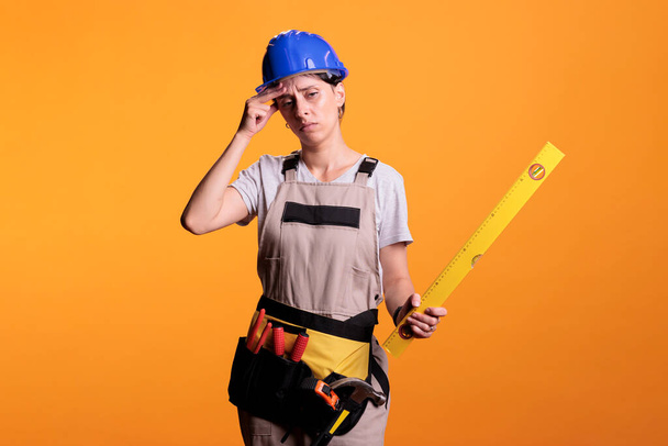 Female constructor dealing with severe headache and holding leveler on camera, rubbing temples. Woman renovation expert with painful migraine dressed with overalls and hardhat in studio. - Фото, изображение