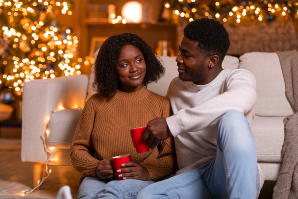 幸せな若い黒人の夫と妻は飲み物のカップで歓声を上げ、休日と余暇を楽しみ、輝くガーランドのあるリビングルームのインテリアで床に座ってください。一緒にお祝い,家で新年, covid-19 - 写真・画像
