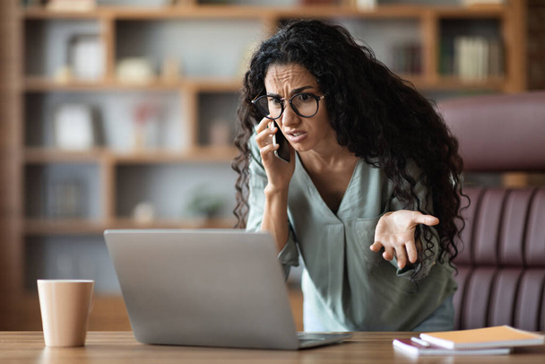 Furious middle eastern young businesswoman wearing eyeglasses having phone conversation with assistant, looking at laptop screen and gesturing, having troubles, office interior, copy space - Photo, image