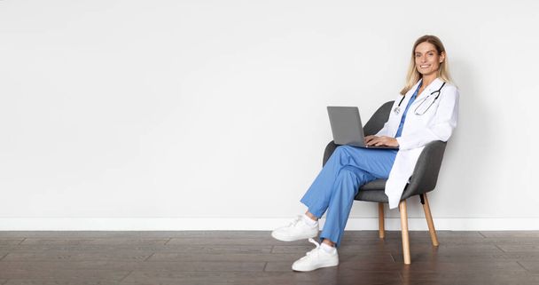 Medical Banner. Lächelnde Ärztin mit Laptop im Sitzen auf Stuhl vor weißer Wand im Krankenhaus, schöne Ärztin in Uniform gibt Online-Beratungen via Computer, Kopierraum - Foto, Bild