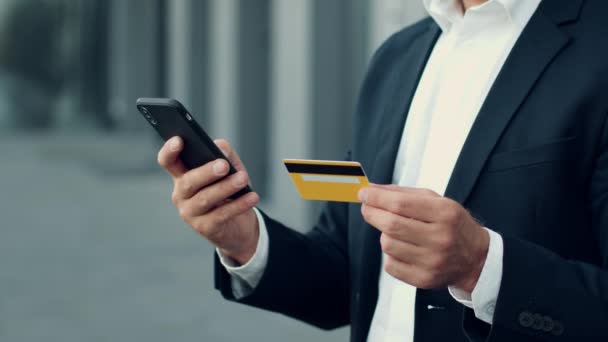 Mobile banking concept. Close up shot of unrecognizable businessman in suit making online financial transaction with credit card and smartphone, standing outdoors, slow motion, empty space - Footage, Video