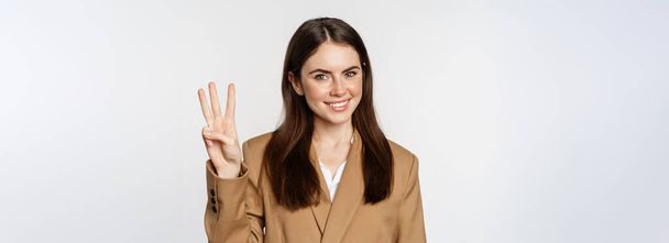 Retrato de la mujer corporativa, vendedora mostrando el número tres dedos y sonriendo, de pie en traje sobre fondo blanco. - Foto, imagen