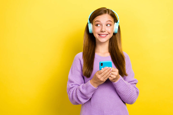 Portrait photo of teenager girl wear warm sweatshirt hold phone wireless headphones listen ebook look empty space isolated on yellow color background. - Фото, изображение