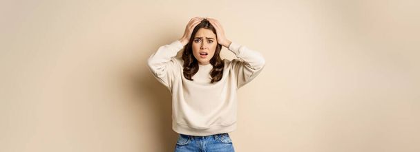 Frustrated and troubled woman facing disaster, holding hands on head in panic, looking anxious, standing over beige background. - Photo, image