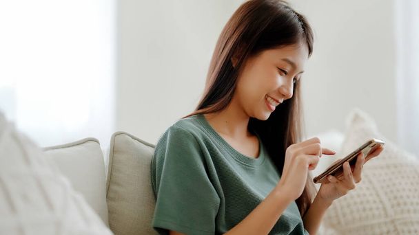 Šťastná mladá Asiatka relaxovat na pohodlném gauči doma textování zpráv na smartphone, usmívající se dívka použít mobilní telefon chatování, procházet bezdrátový internet na gadget, nakupování on-line z domova - Fotografie, Obrázek