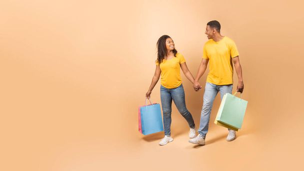 Happy black couple walking with shopping bags and holding hands over peach studio background, panorama with free space. Cheerful man and woman enjoying making purchases together, full length - Photo, Image