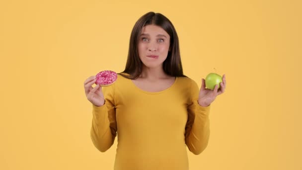 Качели во время беременности. Молодая веселая беременная женщина выбирает между пончиком и свежим яблоком, кусает сладкий десерт, оранжевый фон студии, замедленная съемка, свободное пространство - Кадры, видео