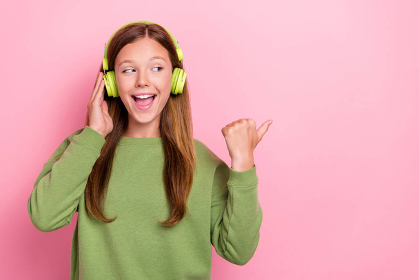 Photo of pretty excite school girl dressed green sweatshirt headphones pointing thumb empty space isolated pink color background. - Zdjęcie, obraz