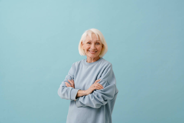 Beautiful smiling senior woman in blue pullover standing with folded arms over isolated blue background - Фото, изображение